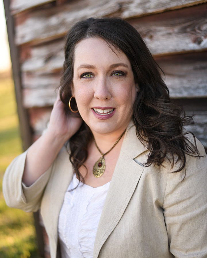 Verity-headshot-WEB-2 Woman with long brown hair, wearing a beige blazer, white blouse, gold necklace, and hoop earring, standing outdoors next to a weathered wooden wall.